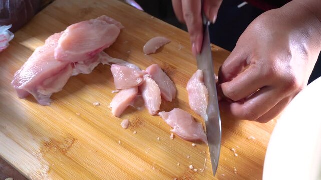 Close-up of human hands slicing raw fresh chicken breast meat with a sharp chef's knife on a wooden cutting board. Perfect for cooking preparation, culinary recipes, food prep, and healthy diet ideas.