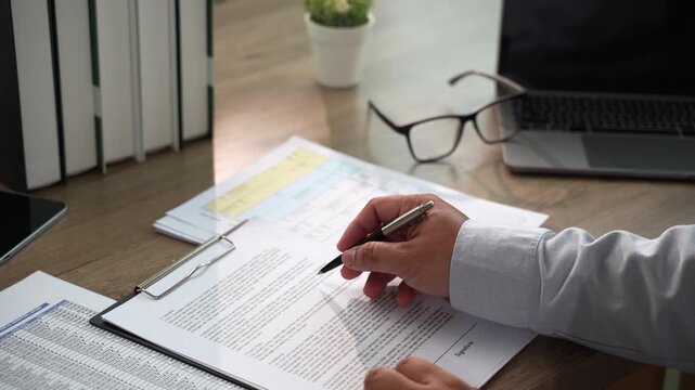 Hand signs a printed contract with pen on clipboard at a wooden desk beside laptop, using daylight workspace for agreement signing, paperwork handling, and contract documentation workflow.