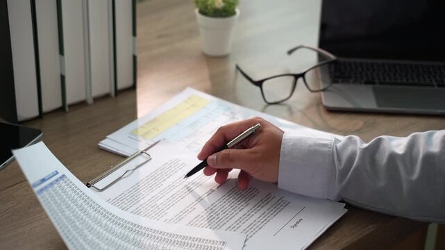 Hand signs a printed contract with pen on clipboard at a wooden desk beside laptop, using daylight workspace for agreement signing, paperwork handling, and contract documentation workflow.