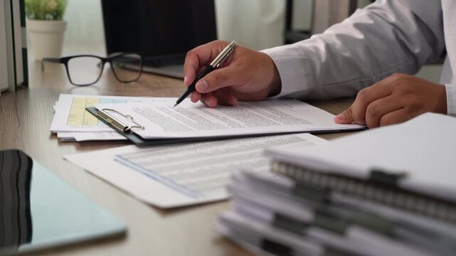 businessman signing over contract papers on clipboard at wooden desk spreadsheet, laptop, glasses, preparing agreement signing, document review, and business paperwork workflow in daylight.