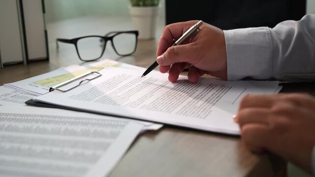 businessman signing over contract papers on clipboard at wooden desk spreadsheet, laptop, glasses, preparing agreement signing, document review, and business paperwork workflow in daylight.