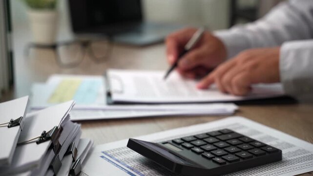 Hand presses calculator beside financial papers while holding pen over documents at desk, checking figures and costs for budget planning, expense calculation, and financial cost review.
