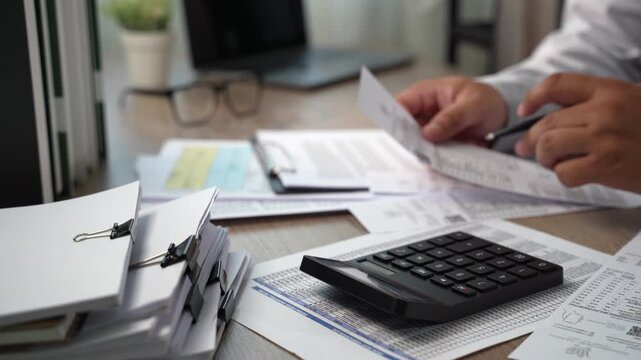 Hand presses calculator beside financial papers while holding pen over documents at desk, checking figures and costs for budget planning, expense calculation, and financial cost review.