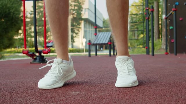 Man in white running sneakers doing warm up jumps on an outdoor sports ground. Athlete doing jumping exercises on an outside gym. Sportsman preparation before a workout in slow motion.