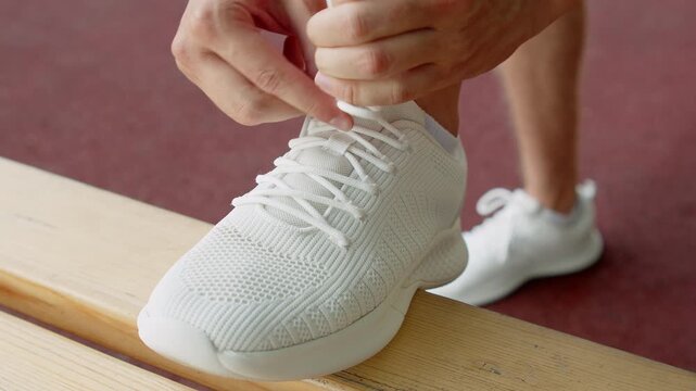 Athlete tying shoelaces before a workout. Tying a white sneaker on a bench close up. Man tying shoelaces on his sport shoes for training outdoors.