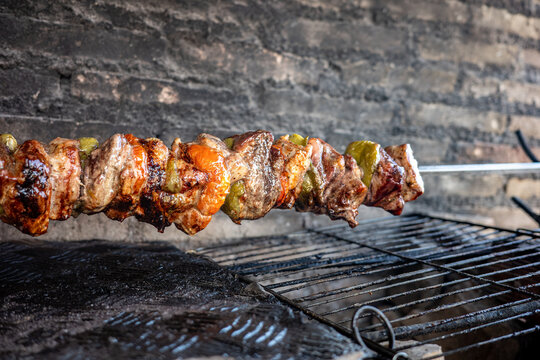 Traditional Greek kontosouvli pork meat chunks marinated with spices and bell peppers roasting slowly on a horizontal metal spit over hot charcoal embers capturing smoke and sizzling details.
