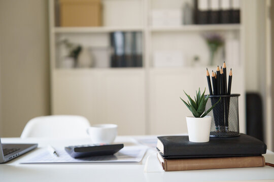 Minimal home office workspace with laptop, calculator, notebooks, pencils, and small plant on a clean white desk.