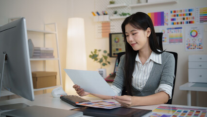 Fototapeta premium Female graphic designer reviewing printed color samples while working at a desk in a modern creative office.