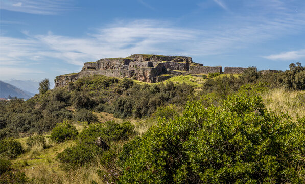 Ancient Inca fortress of Puka Pukara rising above terraced stone walls and lush Andean vegetation with distant mountain scenery under a clear blue sky