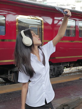 A young Asian woman wearing a university student uniform and over-ear headphones holds up her smartphone to take a selfie while standing on a train platform next to a red passenger train car