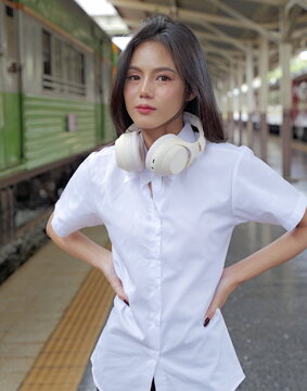 A young Asian woman wearing a university student uniform stands on a train platform with over-ear headphones resting around her neck and hands on her hips, looking directly at the camera