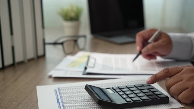 Hand presses calculator beside financial papers while holding pen over documents at desk, checking figures and costs for budget planning, expense calculation, and financial cost review.