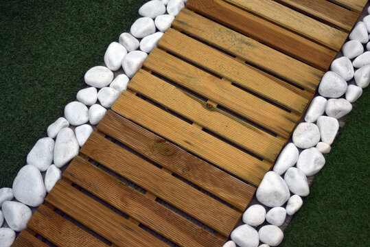 Detail of a garden pathway featuring artificial grass, wooden decking tiles and white pebbles along the border, creating a modern outdoor landscaping design.