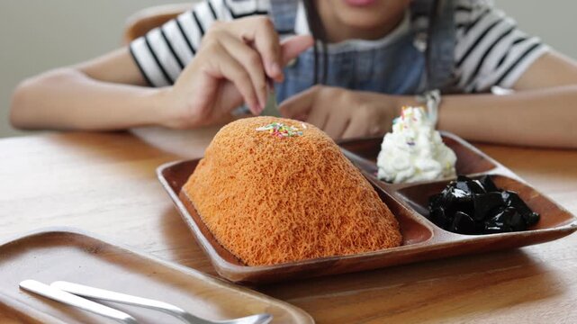 Close-up of young Asian girl using a spoon to eat Thai tea shaved ice bingsu served with whipped cream and grass jelly. Action video footage showcasing sweet dessert enjoyment in a cafe.
