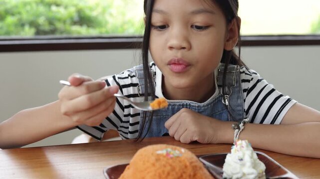 Close-up of young Asian girl holding a spoon, happily eating orange bingsu shaved ice with whipped cream at a wooden cafe table. Natural light video footage of childhood dessert enjoyment.