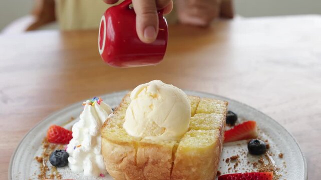 Close-up of hand pouring sweet honey from red pitcher onto vanilla ice cream scope on crisp honey toast. Decorated with strawberries and blueberries. Slow motion video footage for cafe food menu.