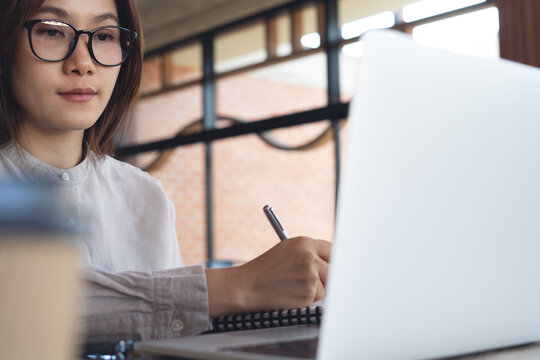 Asian business woman writing in notebook while working remotely on laptop at coffee shop. Freelancer planning work project, online working at cafe, freelance lifestyle concept