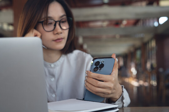 Asian businesswoman using smartphone and working on laptop in cafe modern workspace, remote work, business casual, online business lifestyle, focused multitasking concept.