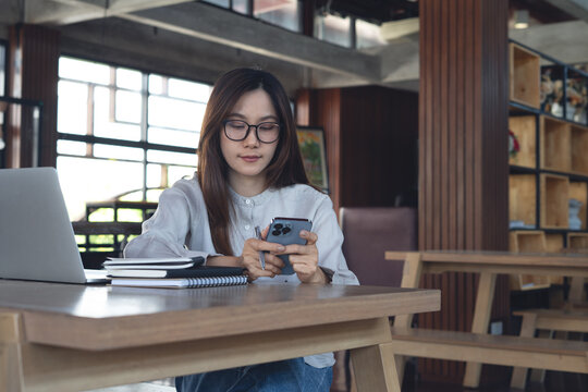 Asian businesswoman using smartphone and working on laptop in cafe modern workspace, remote work, online business lifestyle. Student studying online, e-learning concept
