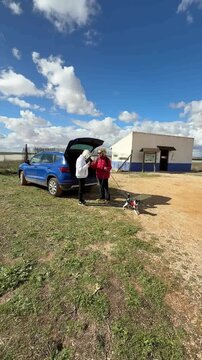 Teenage son checking the hiking route on his smartphone while his mother waits with their small dog beside the car before starting a countryside walk