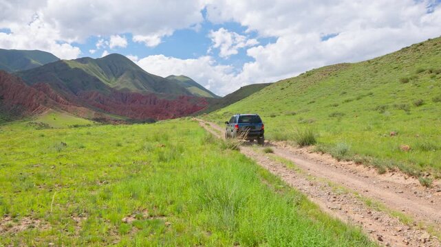 SUV drives along a bumpy dirt road through vast green hills and valleys in Kyrgyzstan. Red rock formations in the background under blue sky with clouds. Scenic off-road landscape with no people.