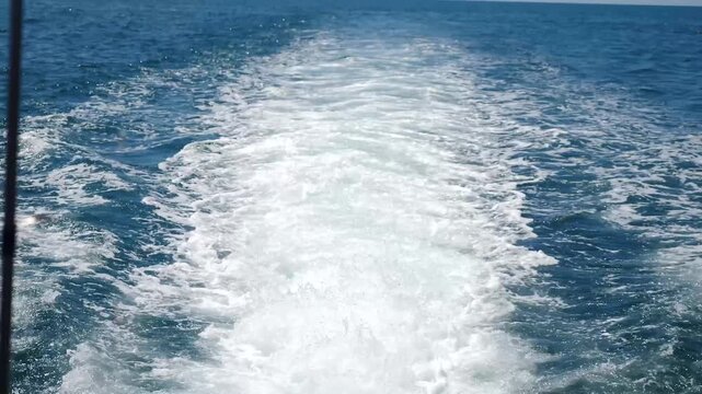 Wide rear view from fast moving ship stern showing powerful white foam wake trail on deep blue ocean surface. High speed maritime vessel navigation creates strong turbulent splash and spray effect. 4k