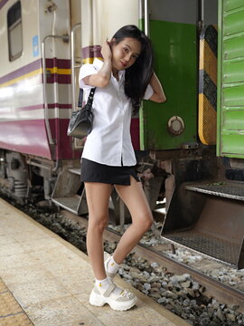 A young Asian woman in a white short-sleeved shirt and black mini skirt stands next to a passenger train car on a station platform