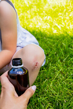Treating child scraped knee wound with antiseptic bottle and cotton pad outdoors.