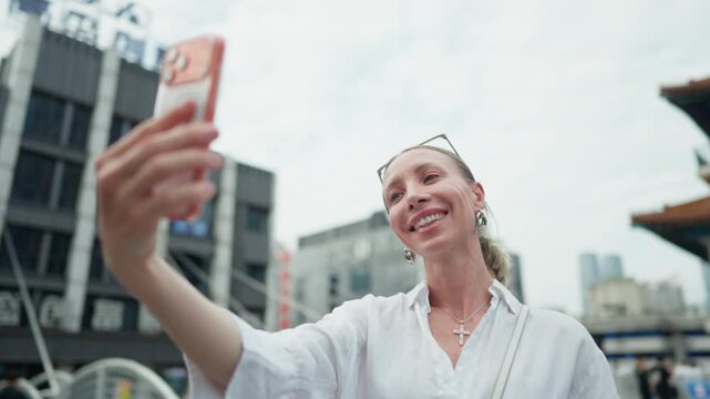Beautiful female traveler taking selfie by mobile phone in downtown in China. Travel in Asia, discover ancient culture and landmarks, admiring architecture of last centuries and modern buildings