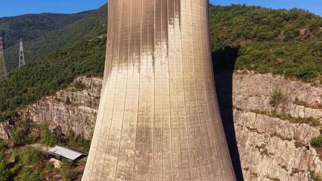 Close aerial view of a weathered concrete cooling tower at a power plant, revealing hyperbolic curve and streaked texture, with rocky gorge, forested mountains and transmission tower below