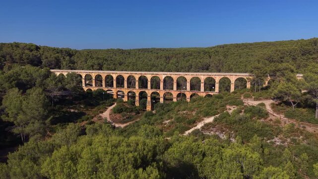 Aerial view of the ancient Roman Pont del Diable aqueduct near Tarragona, Spain, showing its two-tiered stone arches spanning a green wooded ravine under a clear blue sky
