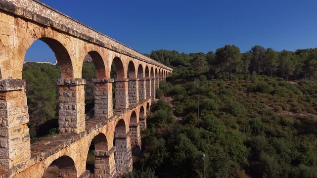 Aerial view along the Pont del Diable Roman aqueduct near Tarragona, Spain, stone arches receding into distance with modern apartment visible through arch, ancient meets modern