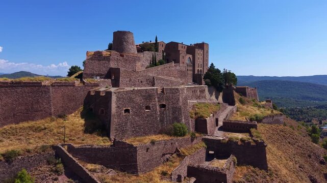 Aerial view of Cardona Castle's medieval stone bastions and cylindrical tower rising above layered fortifications in Catalonia, Spain, with forested hills and blue sky in the background