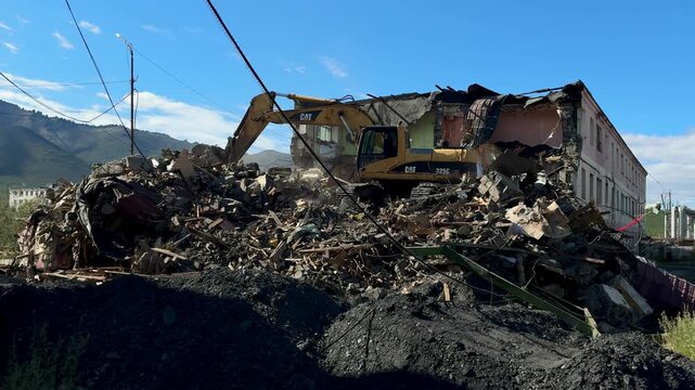 A heavy excavator machine working amidst a massive pile of construction debris and rubble near mountains under a blue sky.