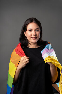 Non binary person on a grey background hugging a LGBTQA+ flag, looking straight into the camera with a smile.