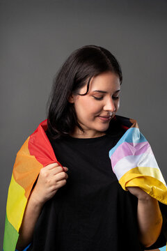 Non binary person on a grey background hugging a LGBTQA+ flag, looking towrads the flag with a smile.