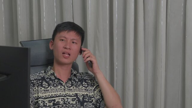 Man talks on phone while seated at desk. He wears patterned shirt, looks engaged. Office setting with monitor and chair. Light gray curtains form background. Imagery captures casual