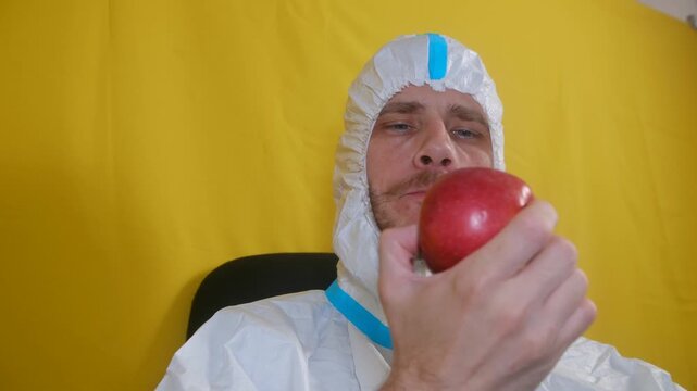 Scientist in white hooded protective suit enjoys healthy snack, eating fresh red apple during his break, against vibrant yellow background. Scientist in protective suit eating a red apple.