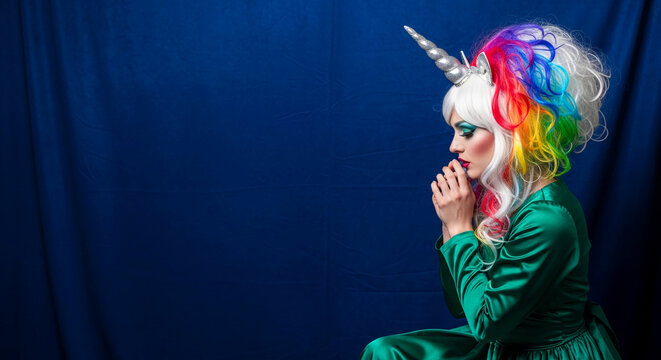 Woman in unicorn horn headband and rainbow wig, with striking makeup, posing against a blue backdrop, symbolizing fantasy and whimsy.