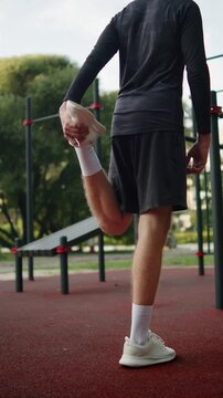 Man doing a standing quadriceps stretch on an outside gym. Man in black athletic clothes pulling his foot to stretch his quadriceps muscle on an outdoor sports ground. Athlete training outdoors.