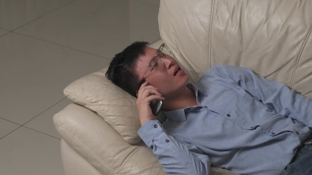 Man rests on sofa, phone to ear, hand raised. He wears glasses and a blue shirt. His expression shows frustration or annoyance. He lies on a cream leather couch