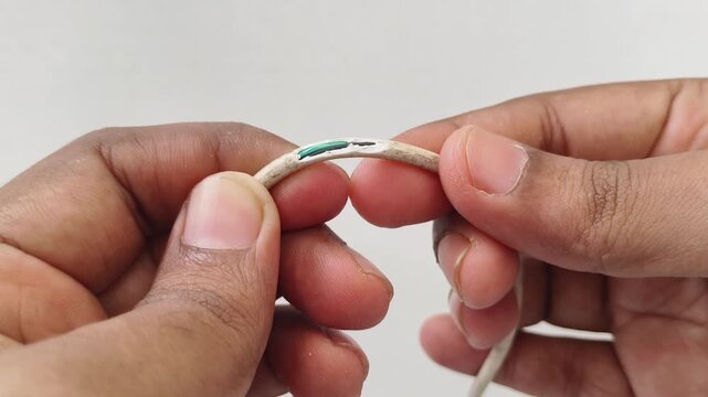 Closeup hands holding and inspecting frayed or damaged wire. Technician or electrician examining cut insulation and exposed copper strands. Old charger cable or electrical power cord wear concept.