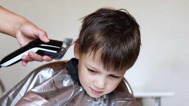 Slow motion close up shows female hand using hair clipper to cut six year old boy hair. Fresh haircut with falling hair is visible. Boy wears barber cape to stay clean.