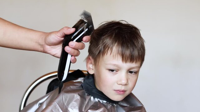 Slow motion close up shows female hand using hair clipper to cut six year old boy hair. Fresh haircut with falling hair is visible. Boy wears barber cape to stay clean.