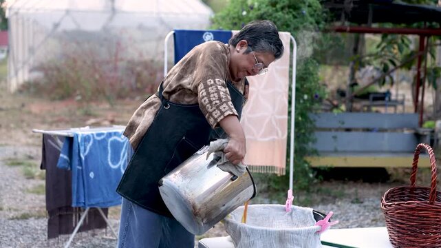 Retired craftswoman pouring natural dye through cloth filter outdoors, handmade textile production, small business process, skilled work, active retirement, productive aging lifestyle.