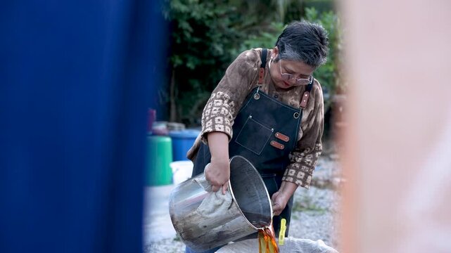 Active senior craftswoman preparing dye solution through cloth filter outdoors, handmade textile workshop, small business process, craft expertise, productive aging lifestyle business.