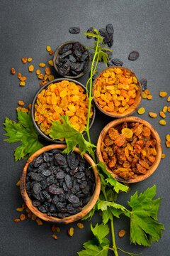 Various bowls filled with golden, brown and dark dried raisins stand on slate background. Fresh green grape leaves garnish colorful composition. Healthy food.