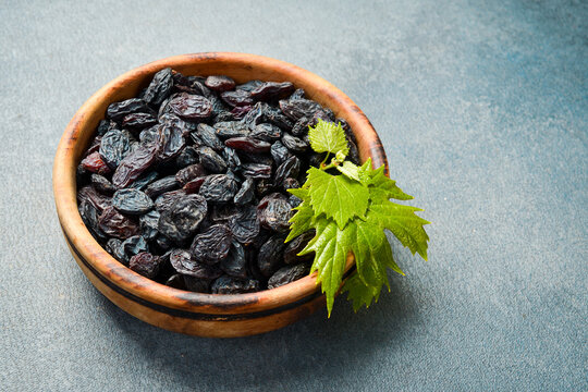 Dark dried raisins fill a round wooden bowl placed on grey slate. Fresh green grape leaves garnish the top of sweet delicious fruits. Healthy food.