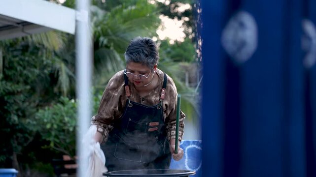 Skilled senior artisan lifting wet fabric from steaming dye bath outdoors, tie dye textile making, craft expertise, small business work, active retirement, productive aging lifestyle.