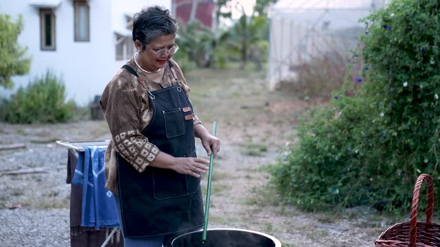 Senior textile entrepreneur stirring dye bath beside finished fabrics outdoors, tie dye craft production, small business activity, creative occupation, productive aging lifestyle business.